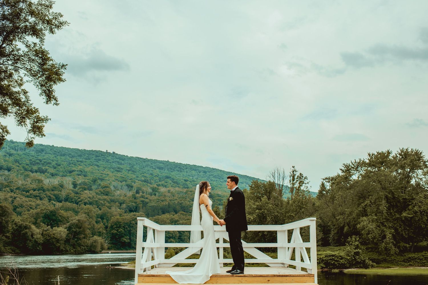 A couple in wedding attire stands on a scenic overlook with rolling hills and trees in the background.
