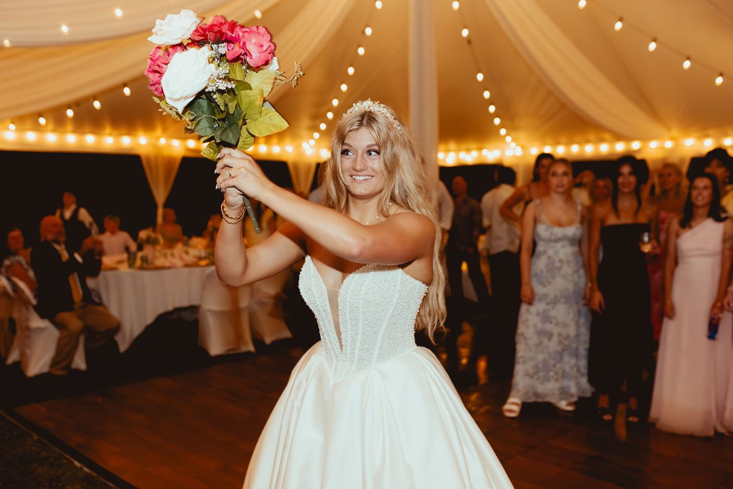 Bride in a white strapless gown tosses her bouquet to guests at a tented wedding reception.