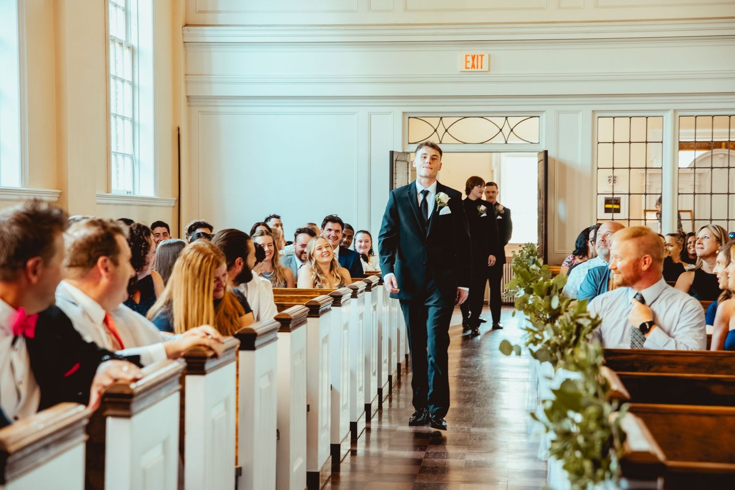 Wedding ceremony in a traditional church with guests seated in wooden pews decorated with greenery along the aisle.
