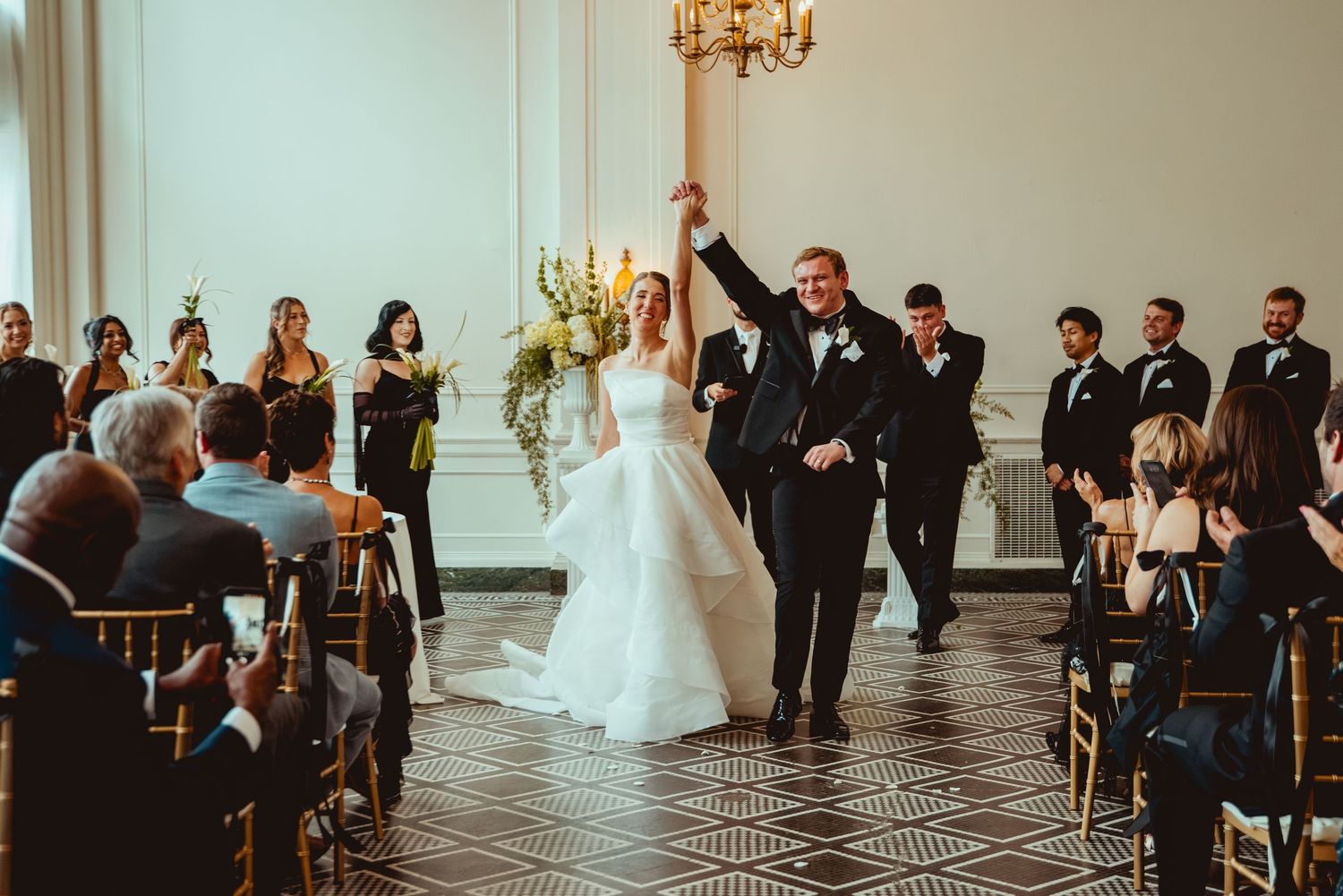 Wedding ceremony in an elegant ballroom with guests seated in rows and a chandelier overhead.