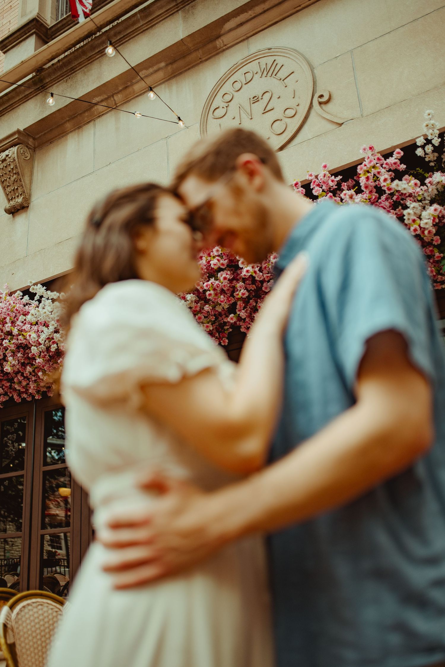 A couple embraces and kisses outside a flower-adorned building with a Goodwill No.2 sign in soft focus.
