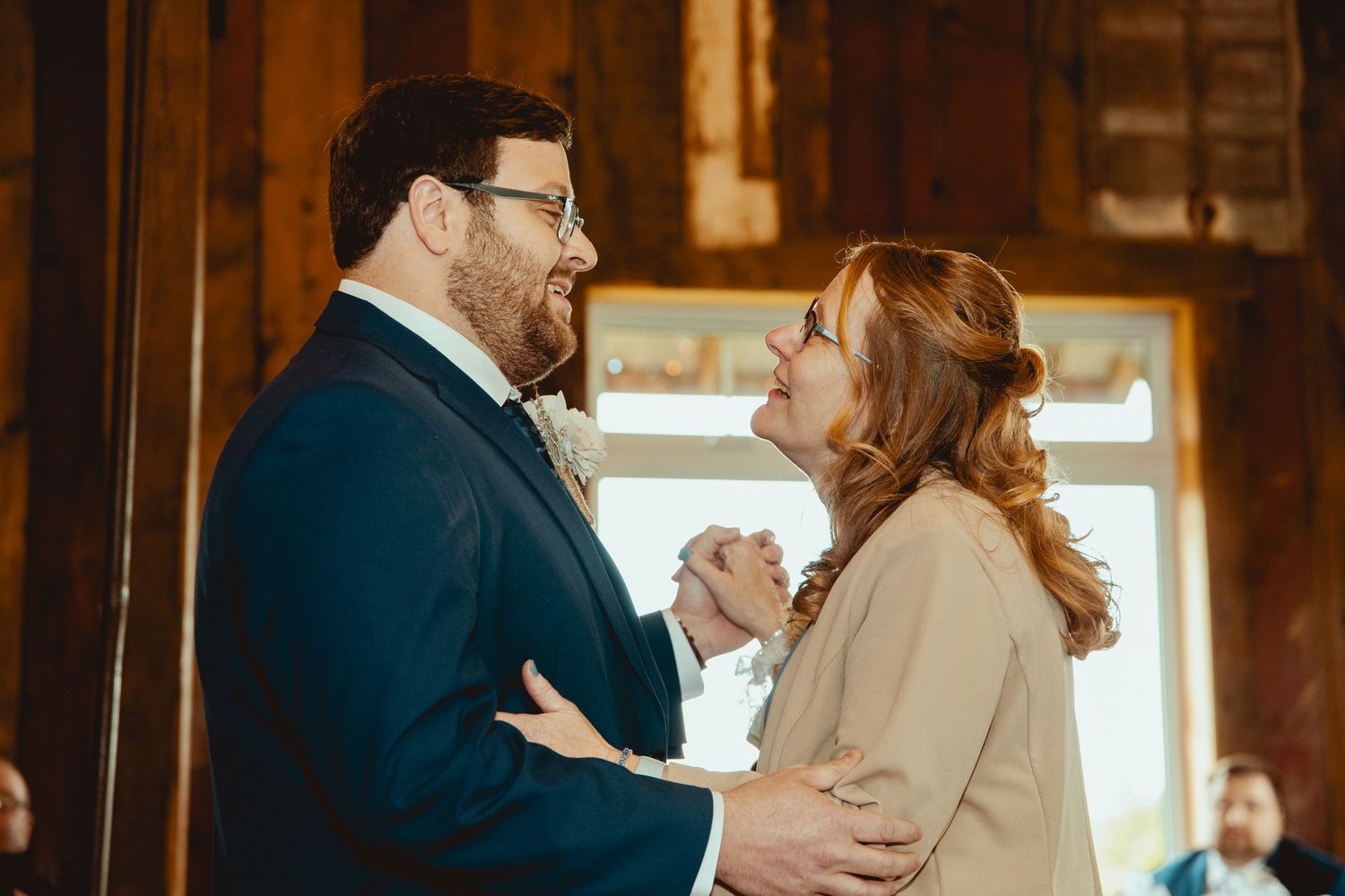 Groom in navy suit sharing emotional mother son dance in warmly lit rustic wedding venue.