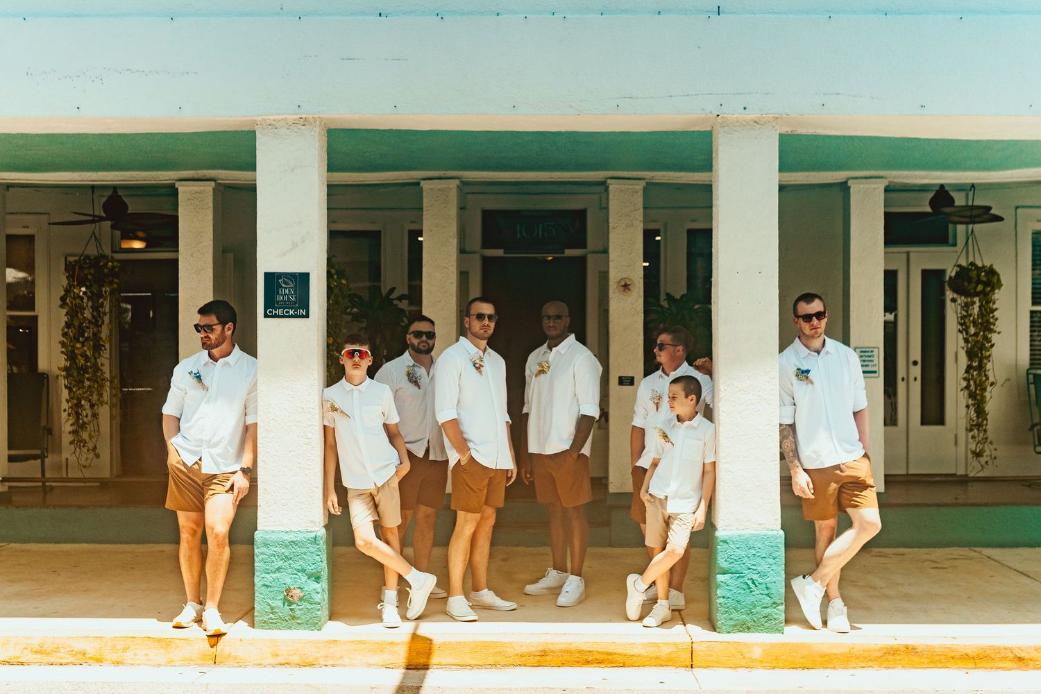 Groomsmen in matching white shirts and tan shorts pose together outside a tropical venue.
