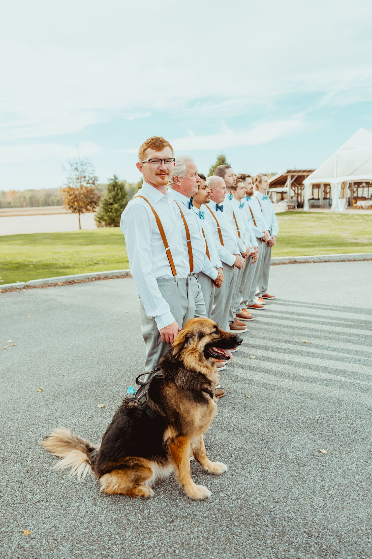 A German Shepherd dog sits on pavement while groomsmen in light blue shirts and suspenders stand in line at a wedding.