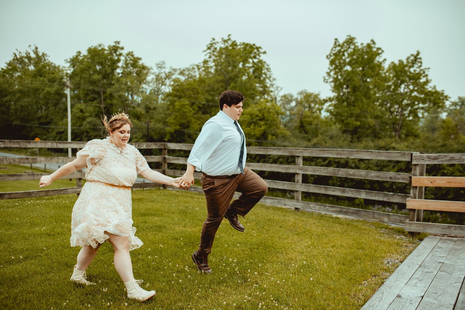 A playful couple jumps and dances on green grass with a wooden fence in the background.