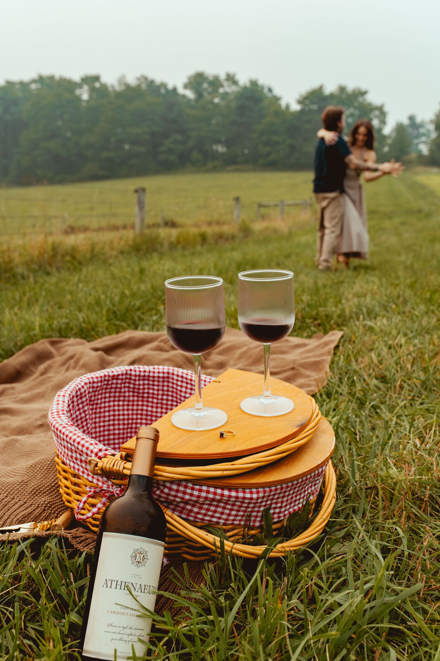 Romantic picnic scene with red wine glasses, wicker basket, and couple dancing in green meadow background.