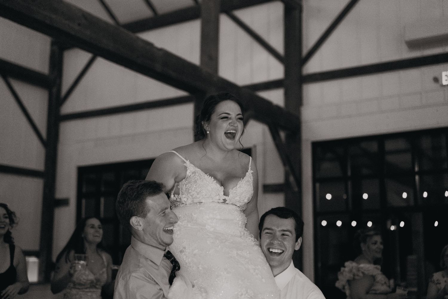 Joyful bride laughs while being lifted by groomsmen during black and white reception celebration.