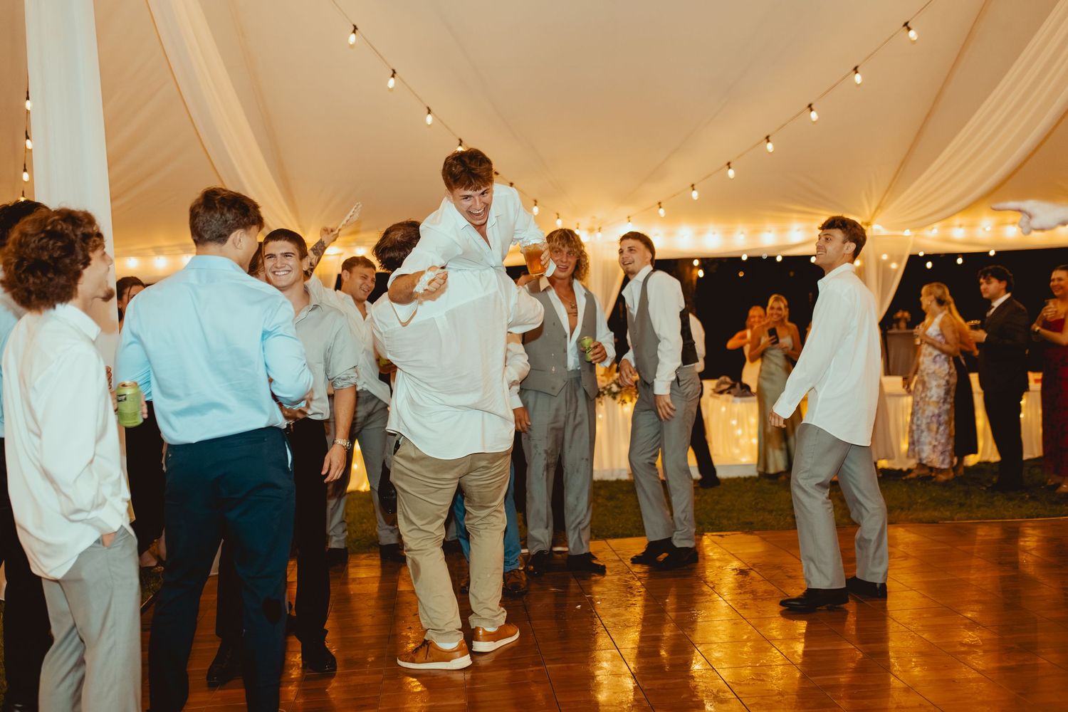 Groomsmen lifting the groom in celebration on the dance floor at a tented wedding reception.