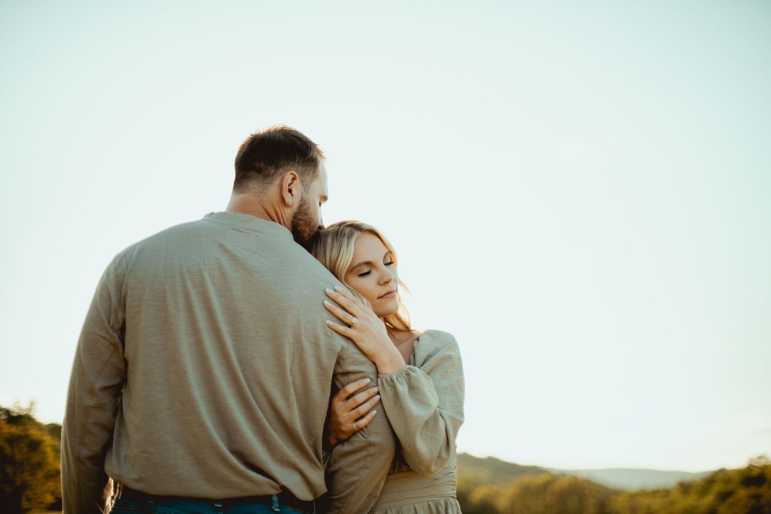 A couple embraces tenderly in a field during golden hour as sunlight illuminates their silhouettes.
