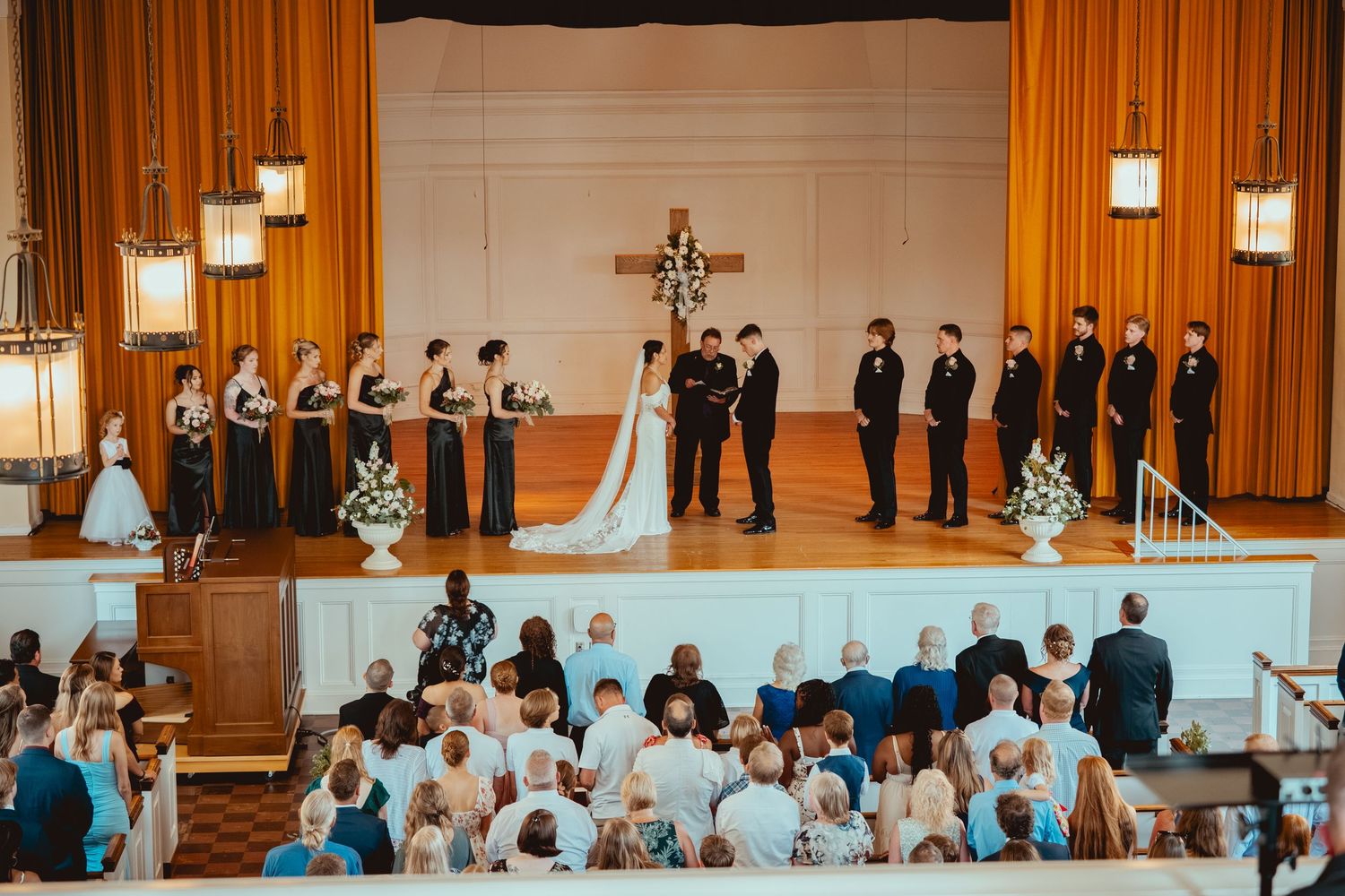 Wedding ceremony taking place on a church altar with orange curtains and hanging pendant lights in the background.