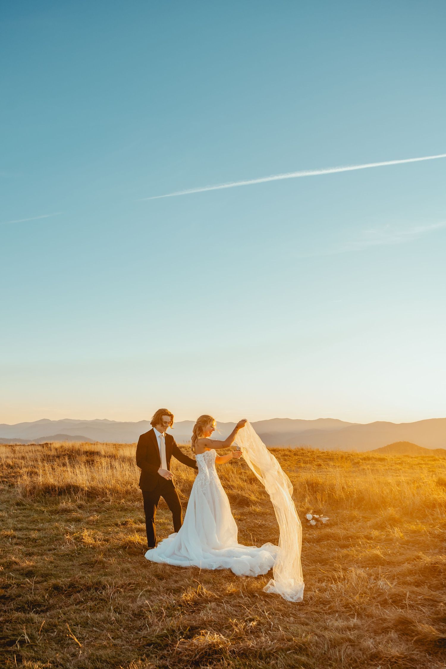 A wedding couple poses against a stunning mountain backdrop during sunset with a flowing veil.