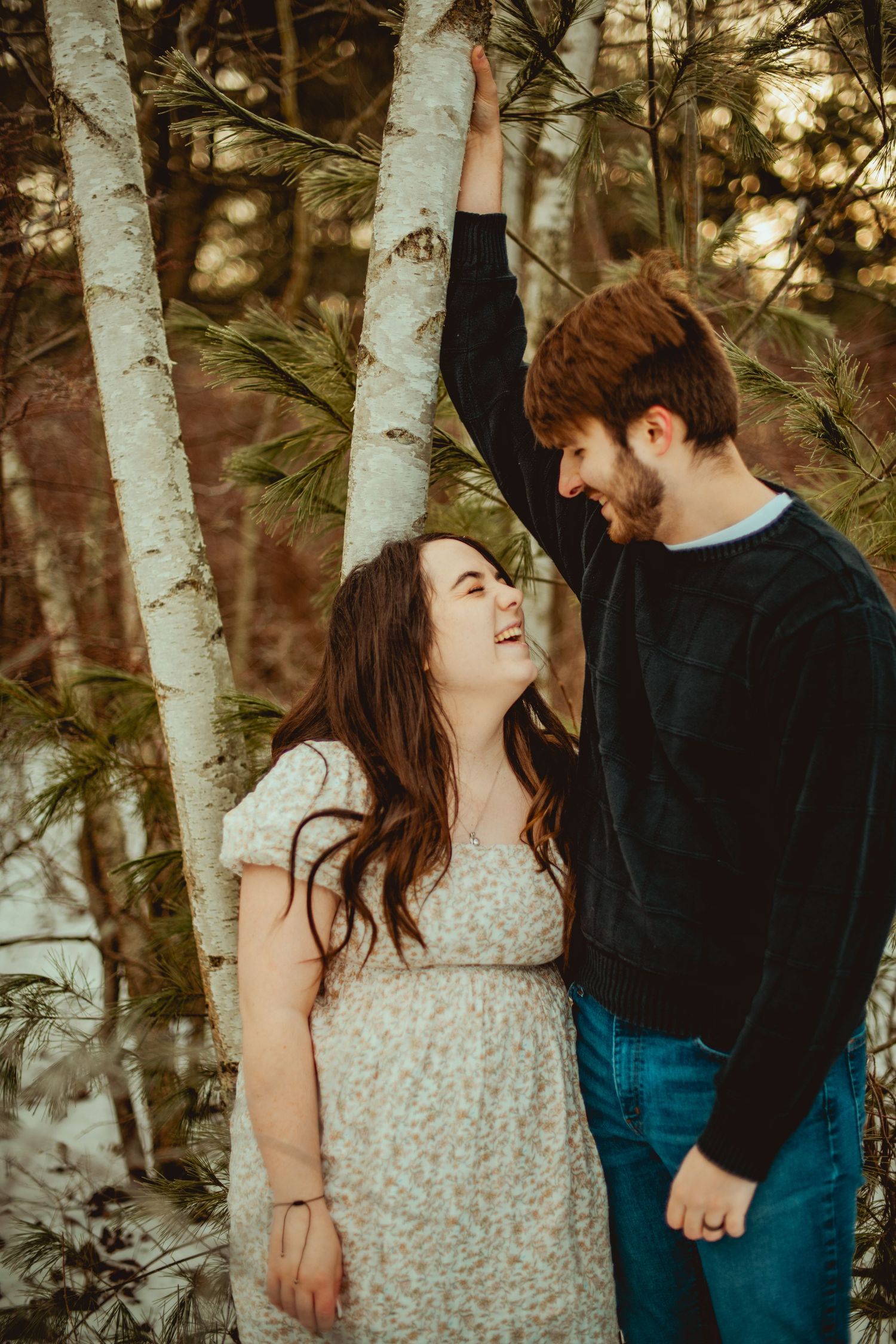 A couple shares a joyful moment between birch trees in a forest setting.