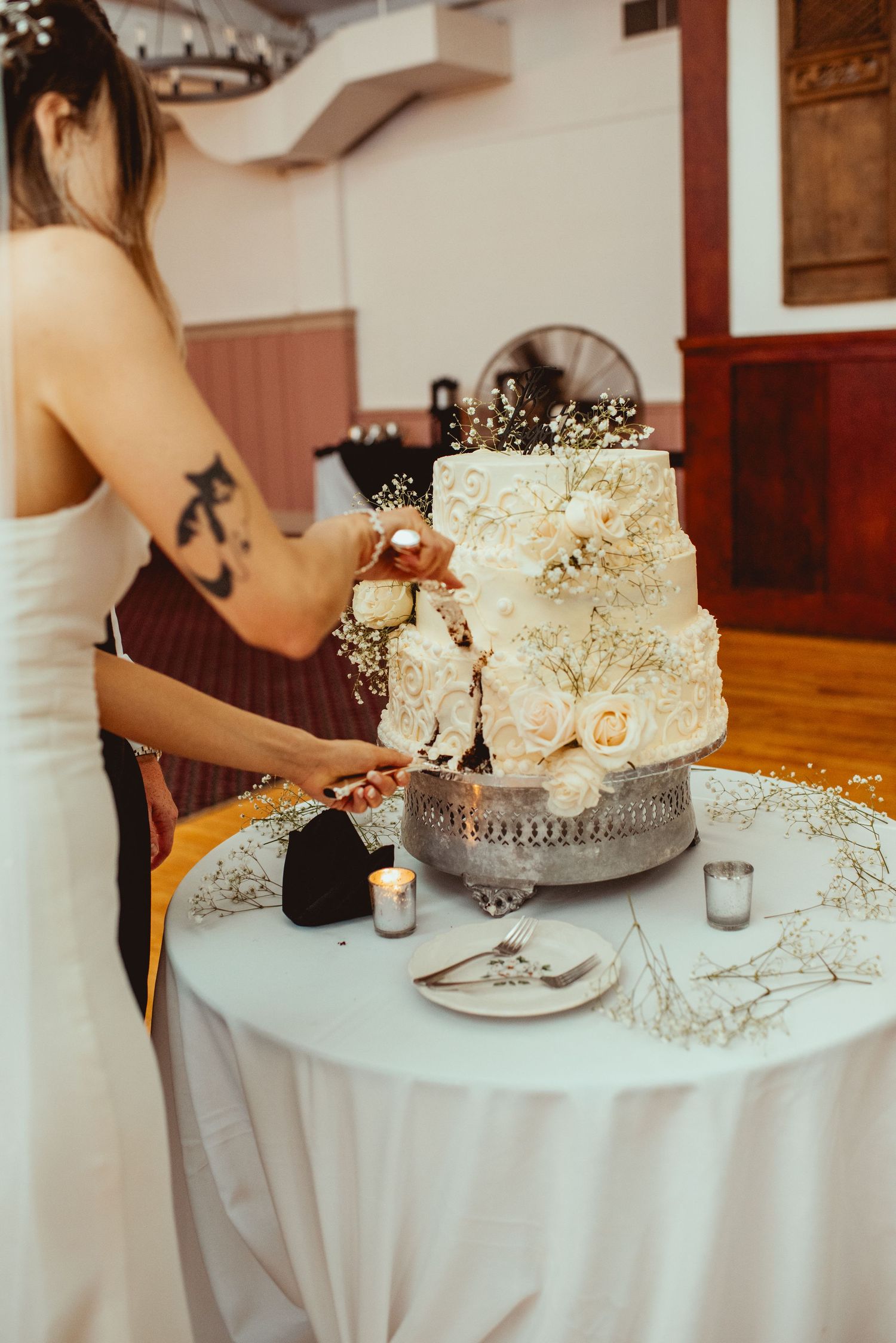 White tiered wedding cake with delicate floral decorations on round table with place settings.