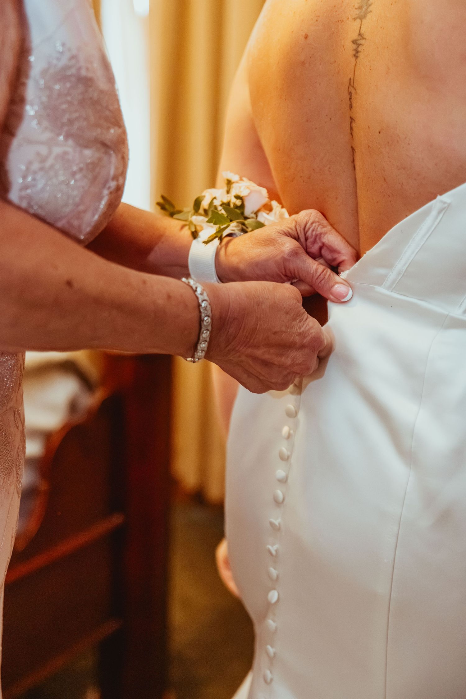 Helper fastening row of buttons on back of white wedding dress on bride.