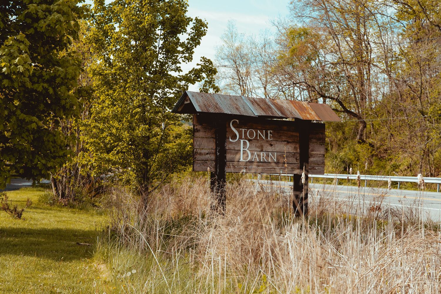 Rustic wooden barn sign reading Stone Barn surrounded by autumn trees and tall grass along a country road.