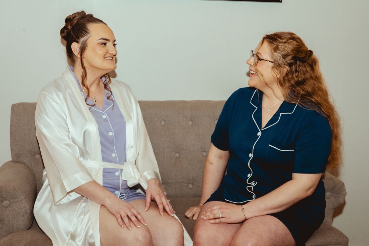 Two women in pajamas sitting on a couch smiling at each other during bridal getting ready preparations.