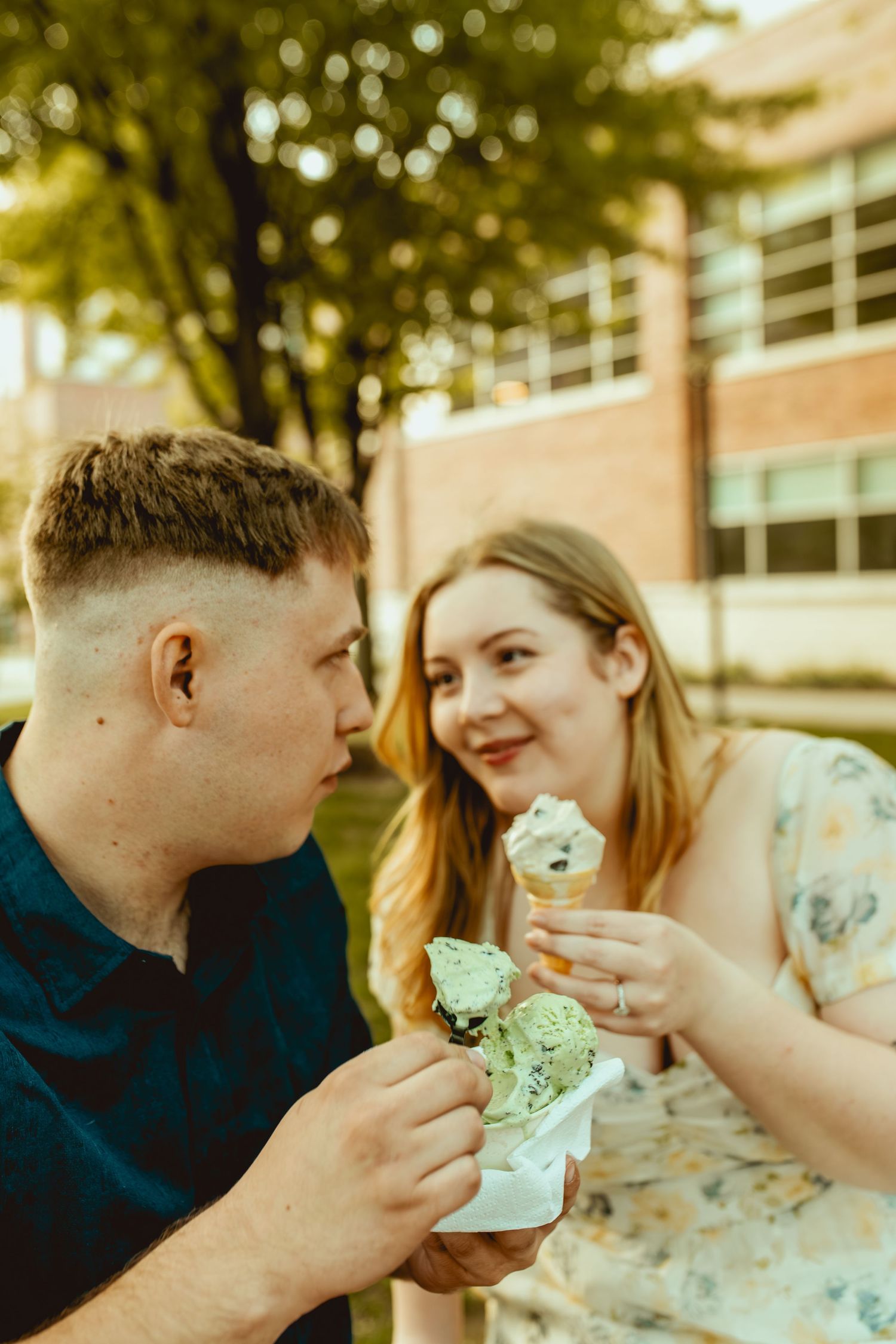 A young couple shares ice cream cones outdoors, smiling at each other near a brick building with green trees.
