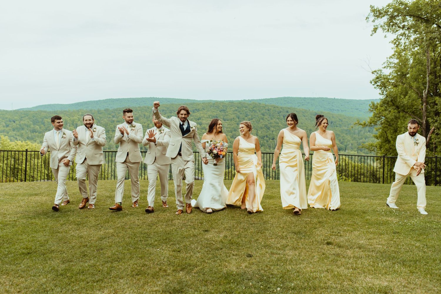 Wedding party walks together on green lawn with scenic mountain views, bride in white gown.