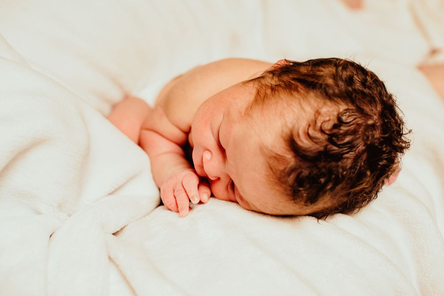 Peaceful newborn baby with dark curly hair sleeping on white blanket in soft lighting.