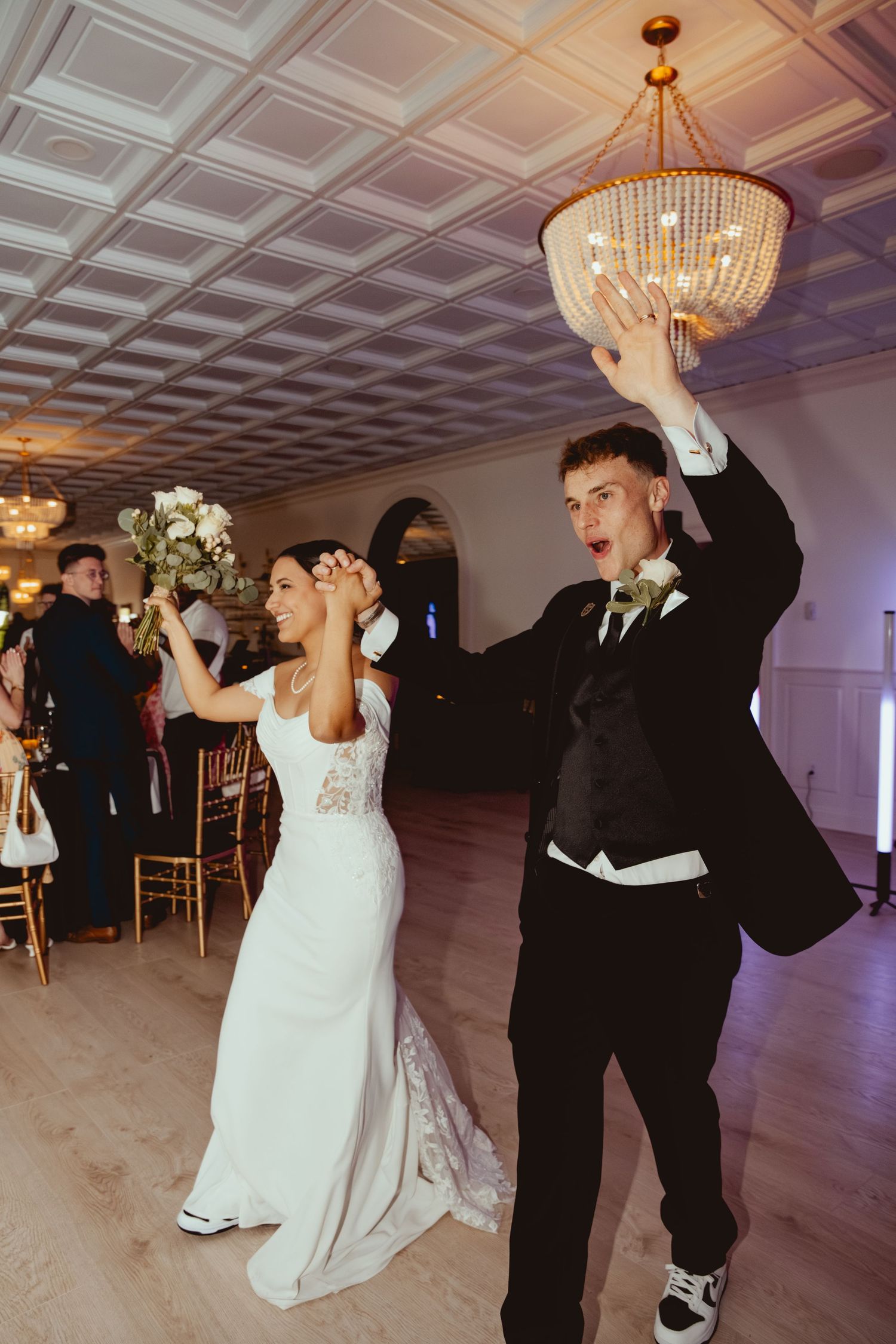 A couple dances joyfully in an elegant ballroom with ornate chandeliers and a coffered ceiling.
