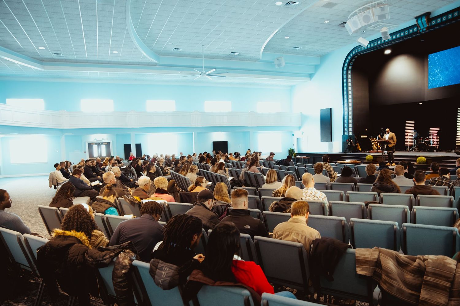 A large audience sits in rows of blue chairs facing a stage during a conference or presentation in an indoor venue.