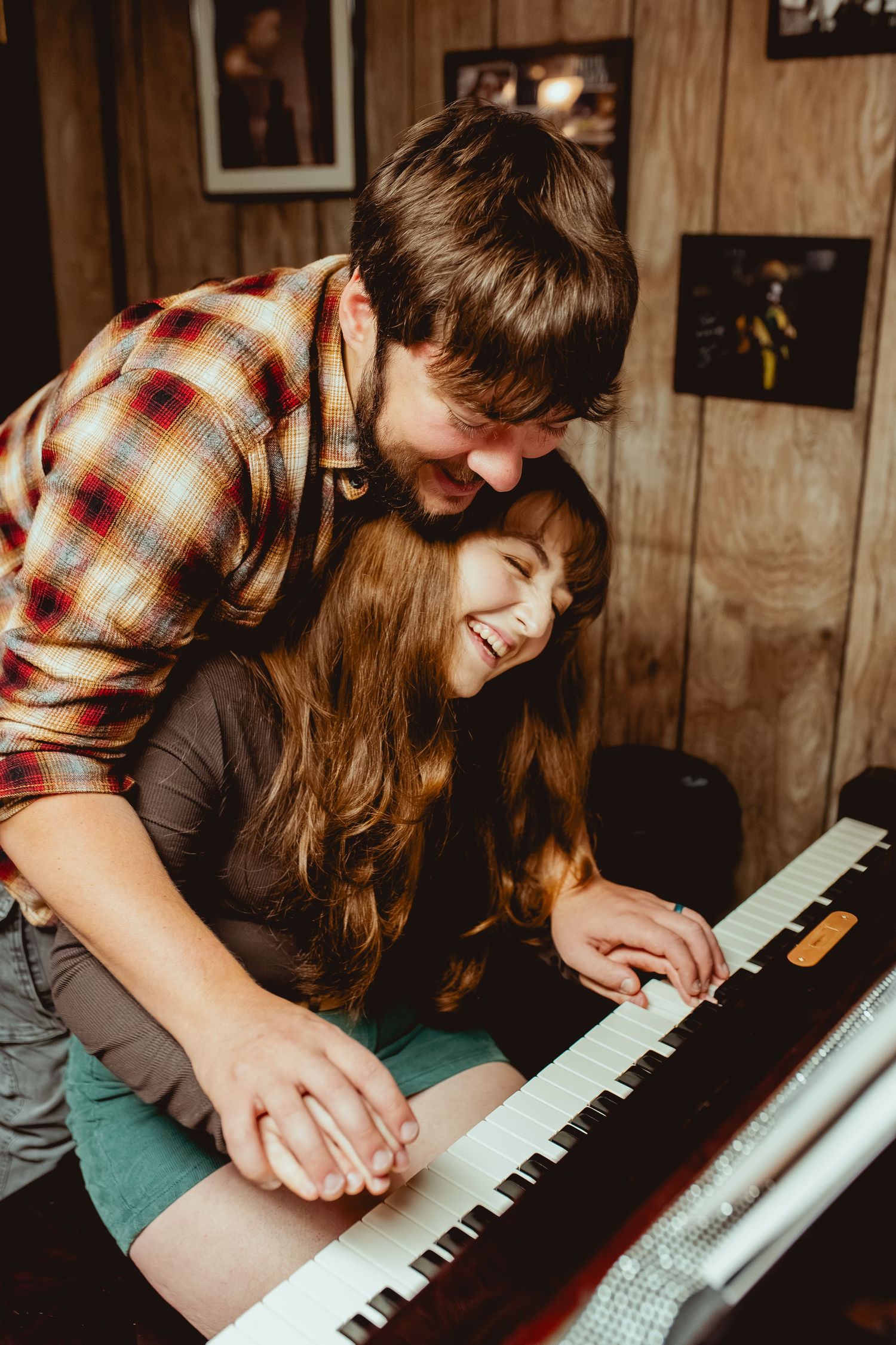 Two people share a playful moment while sitting together at a piano keyboard in a rustic wood-paneled room.