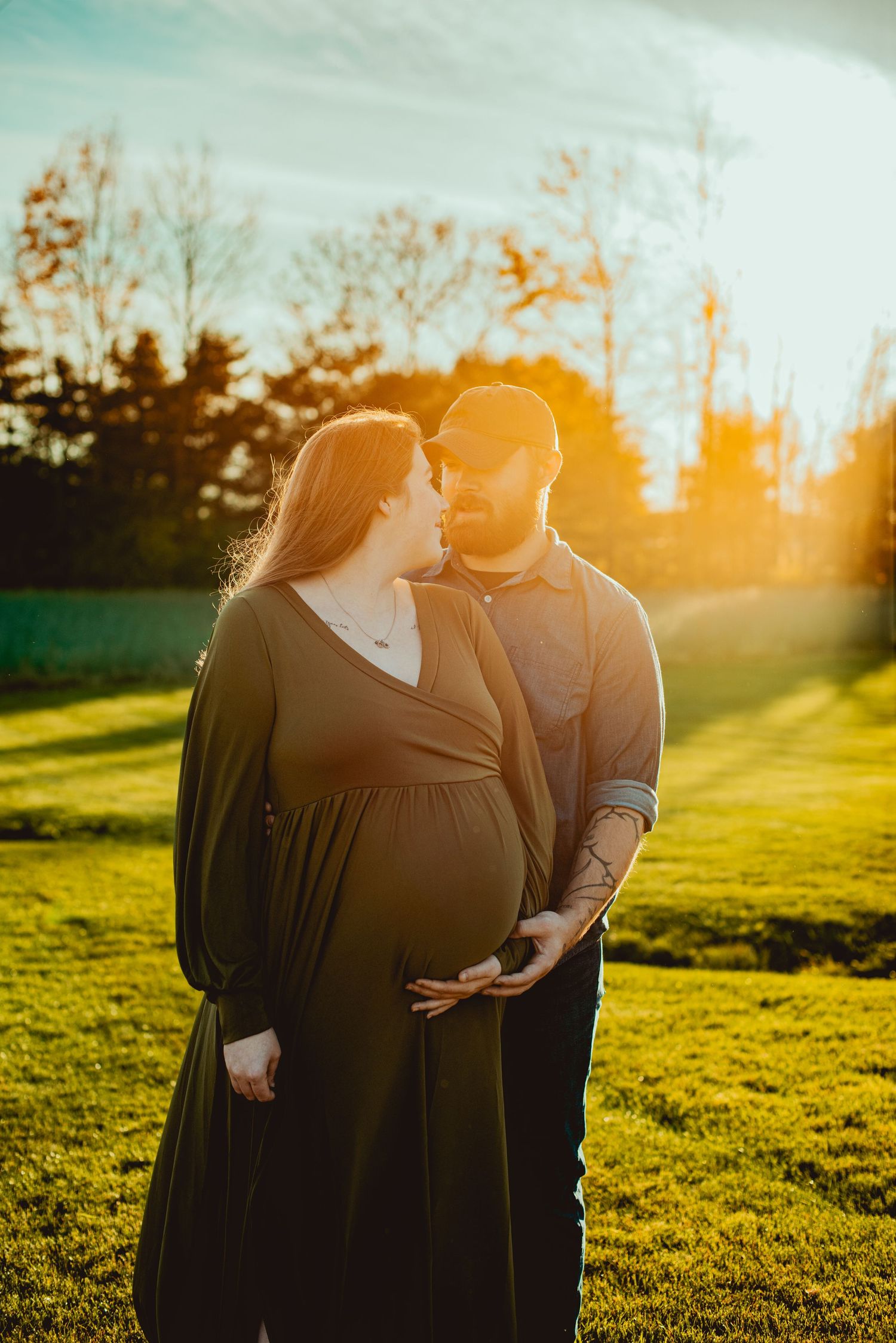 A couple embraces in a grassy field during golden hour sunset as warm sunlight illuminates the scene.