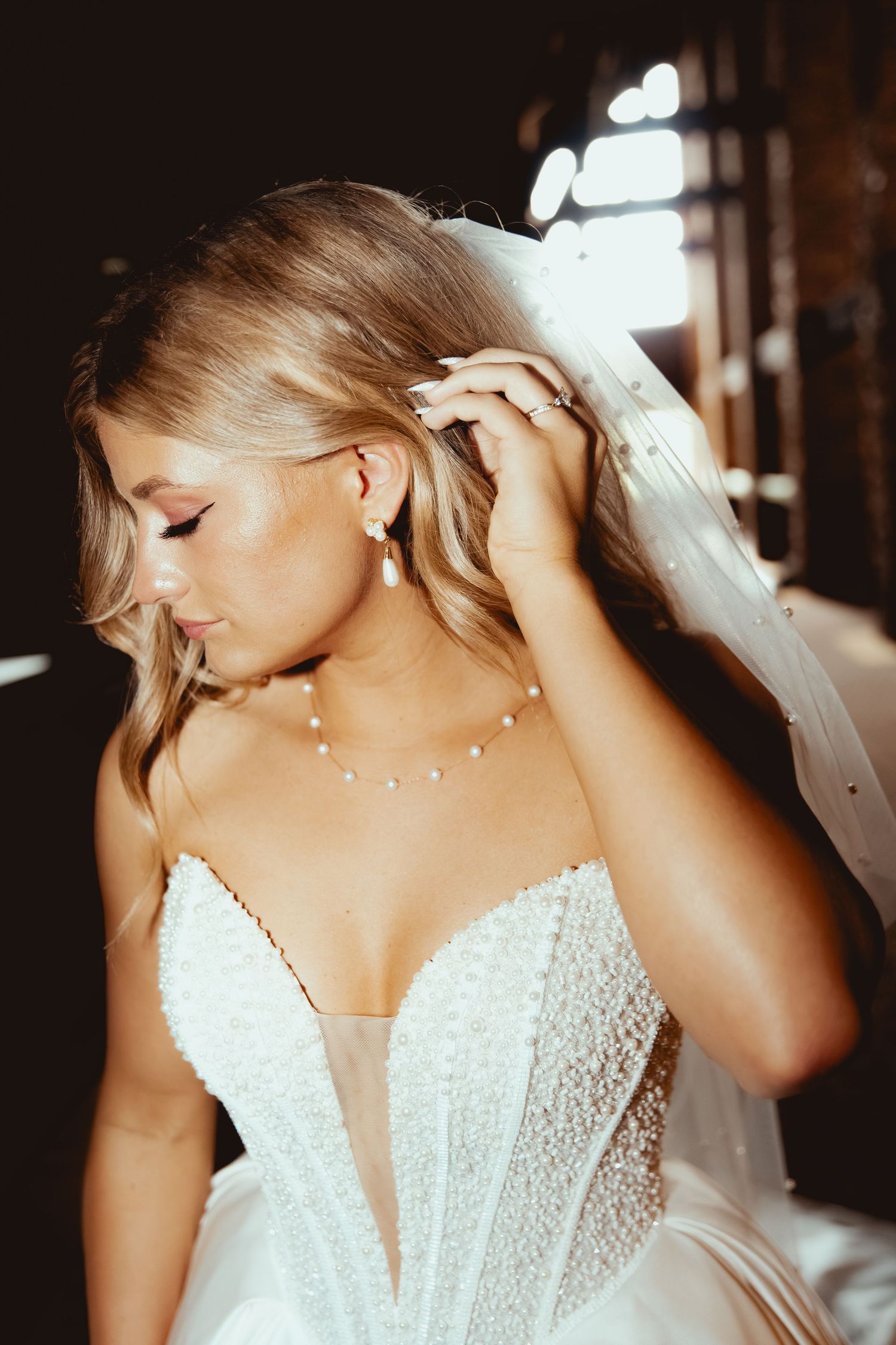 Elegant bride in pearl-embellished wedding gown adjusts her veil with delicate jewelry in soft light.