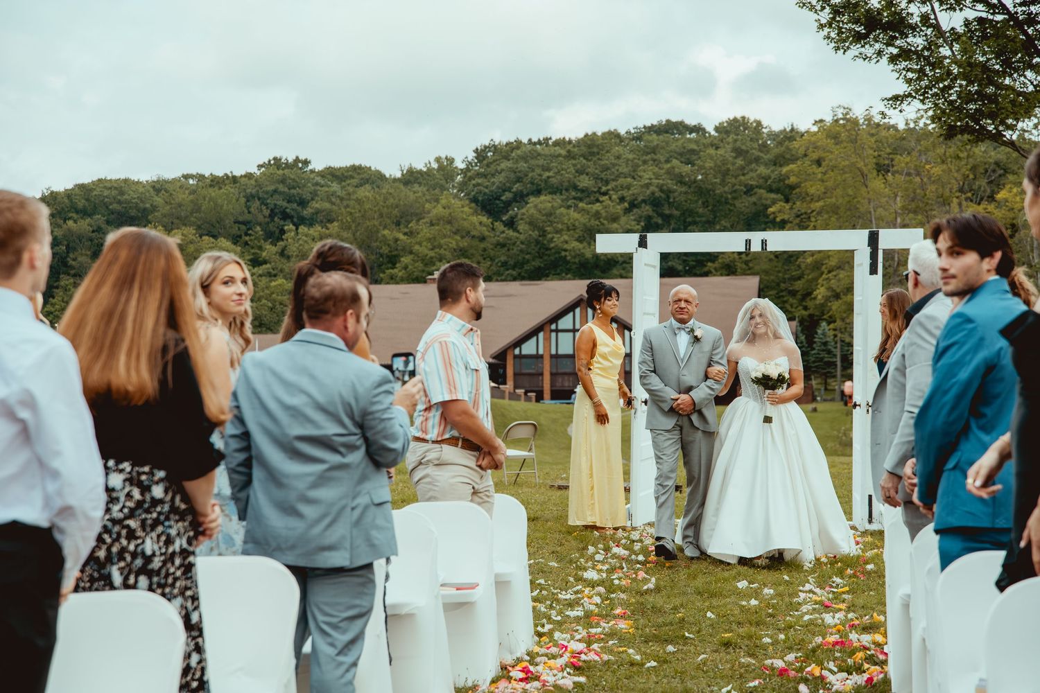 Outdoor wedding ceremony on green lawn with guests standing along aisle, bride in white gown, white arch, rustic venue backdrop.