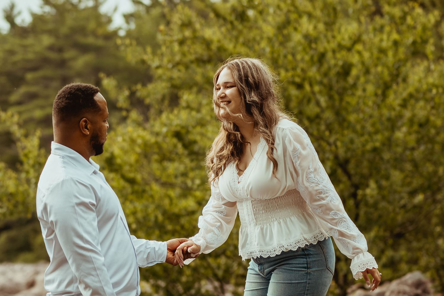 A couple holds hands and smiles at each other outdoors, surrounded by lush green trees.