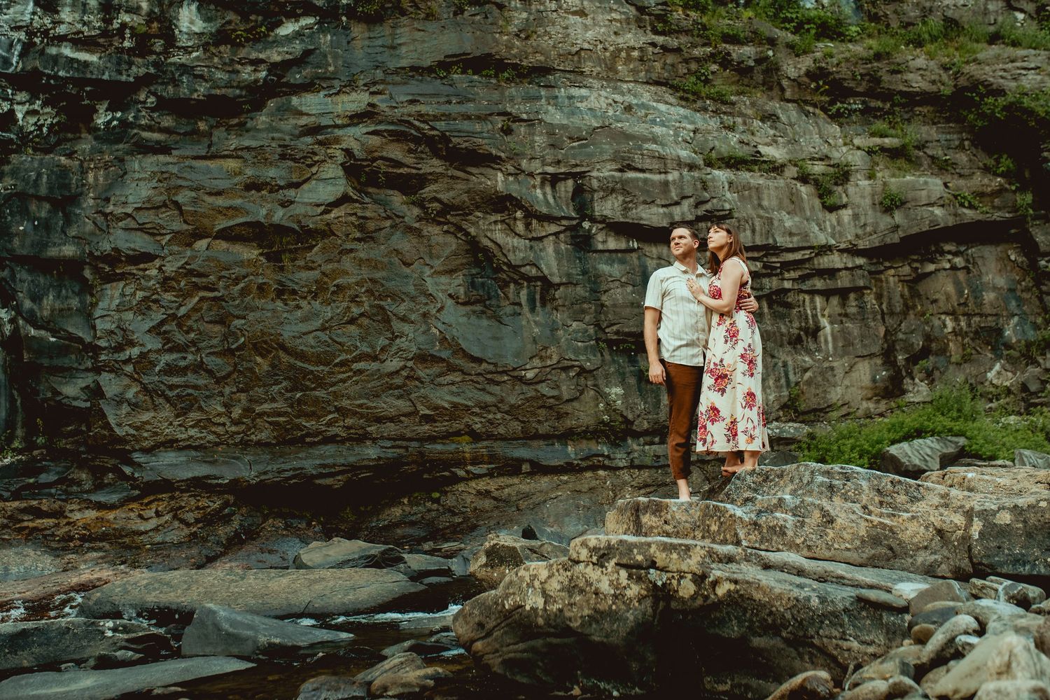 Couple standing on rocks near a stream with a dramatic stone cliff backdrop in a romantic outdoor engagement photo.