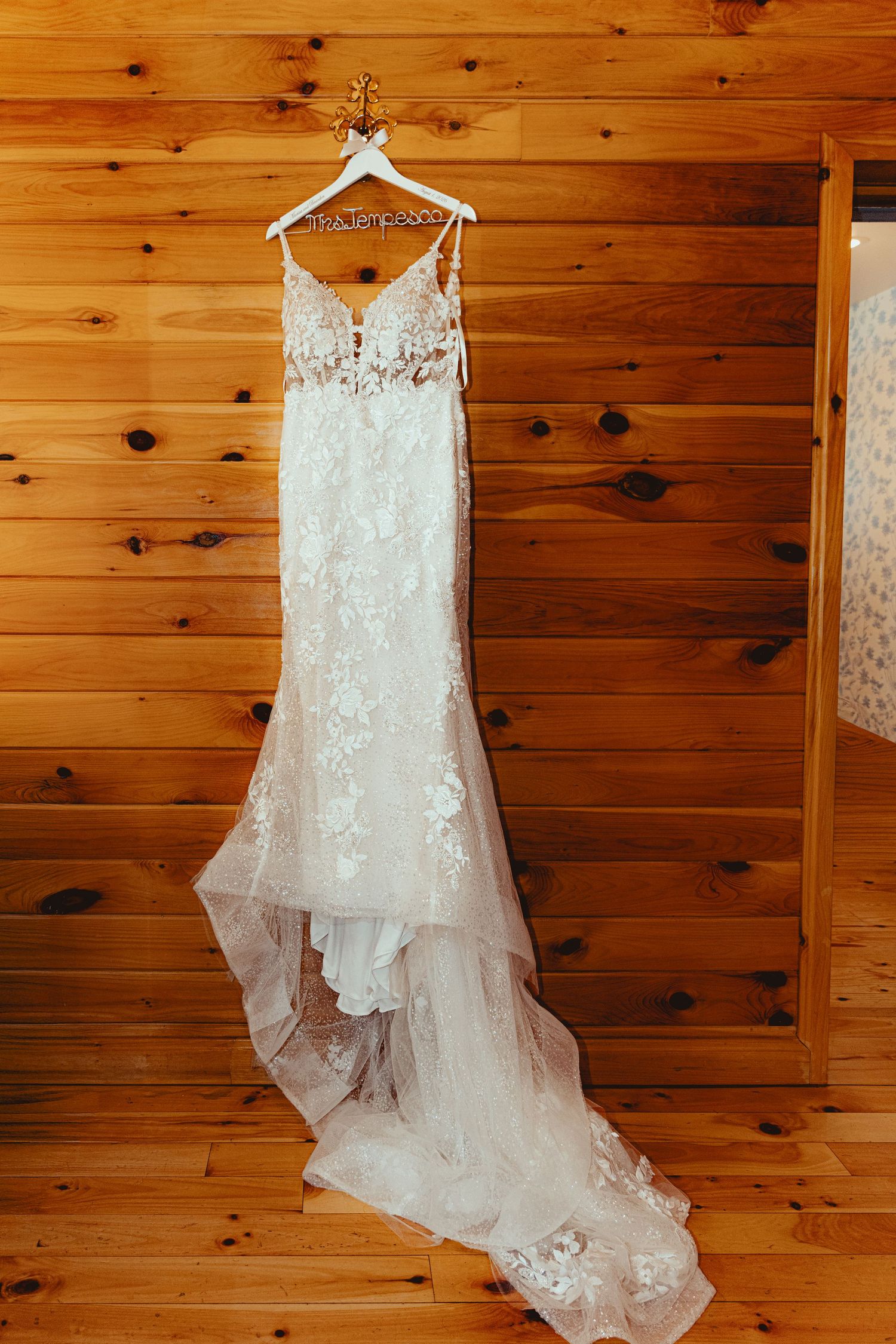 Elegant lace wedding dress with floral details and train hanging on a white hanger against a rustic wood wall.