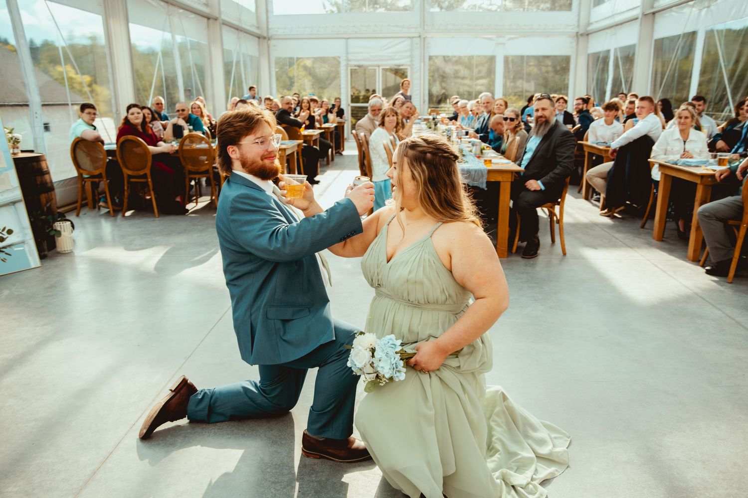 Wedding reception dance party inside elegant glass-roofed venue with wooden tables.