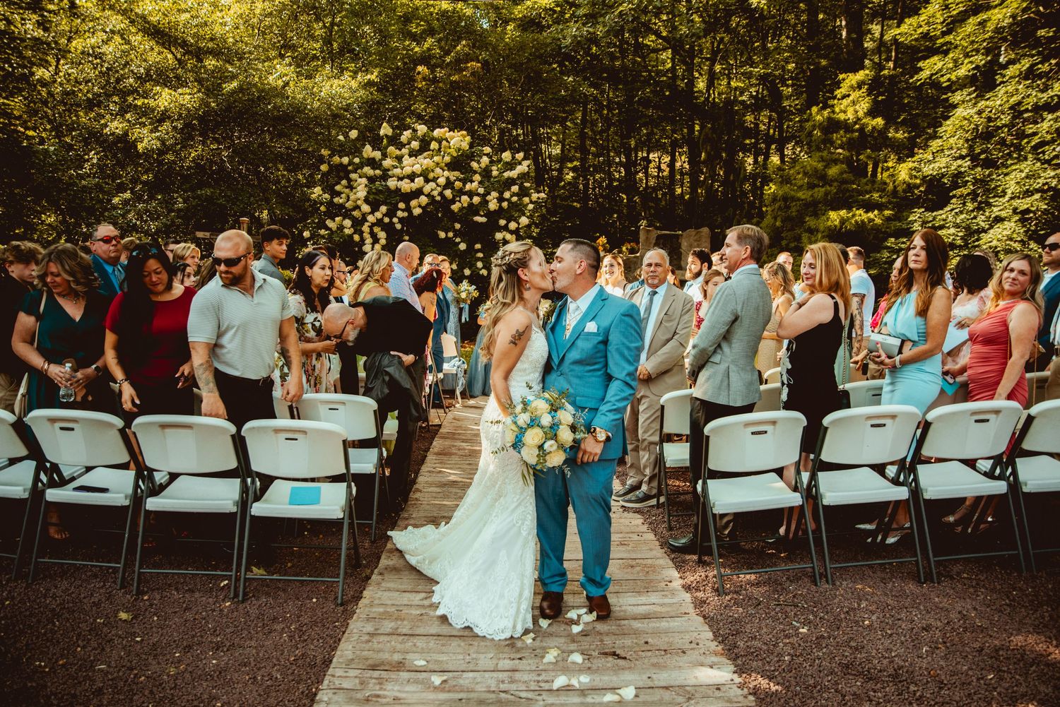 Outdoor wedding ceremony with guests seated on white chairs surrounded by greenery.