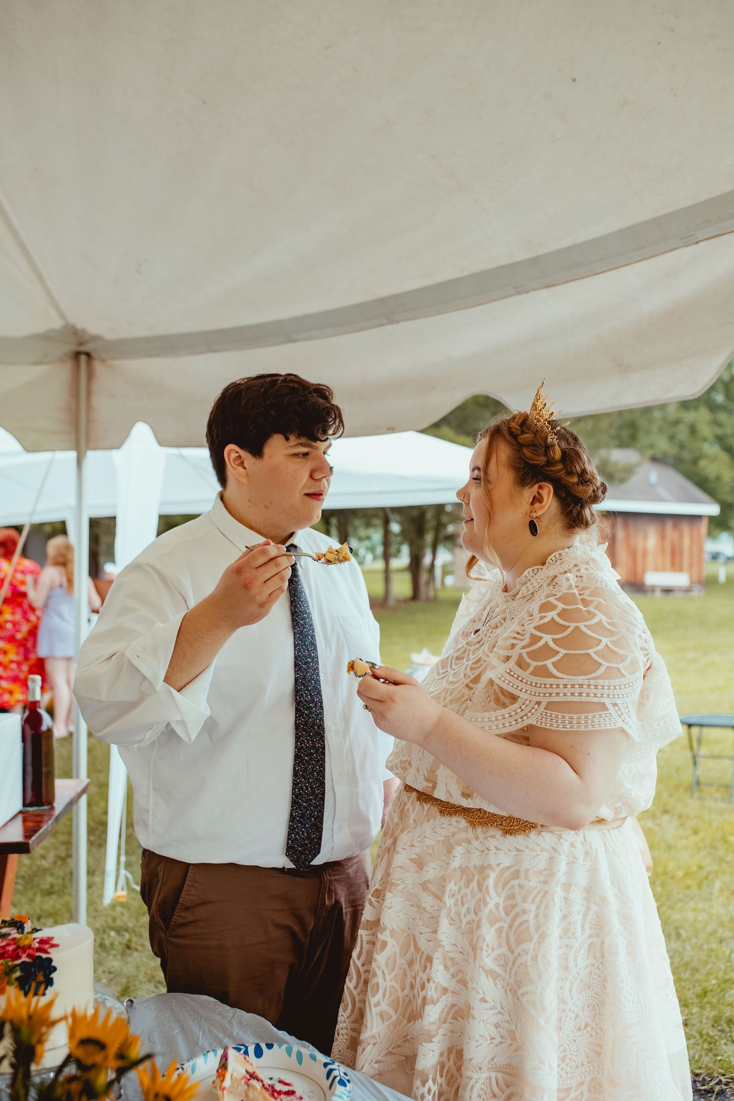 A couple in wedding attire feeds each other cake under a white tent at an outdoor reception.