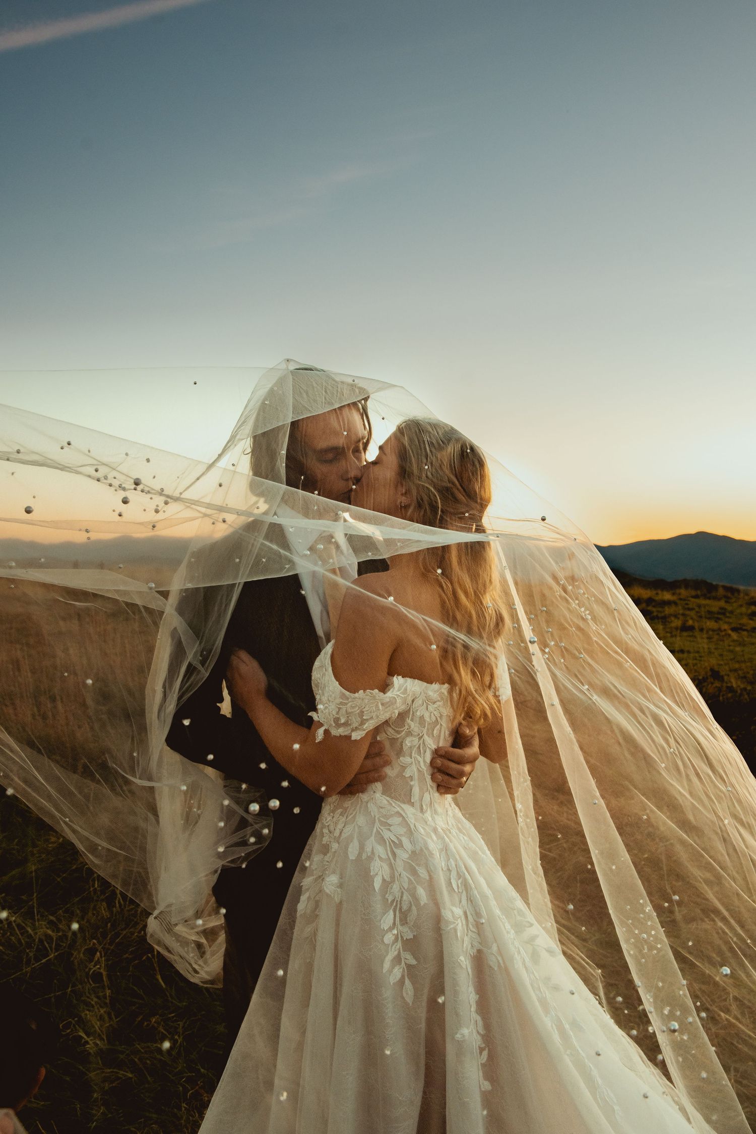 Wedding couple sharing romantic moment under flowing veil with mountain vista backdrop.