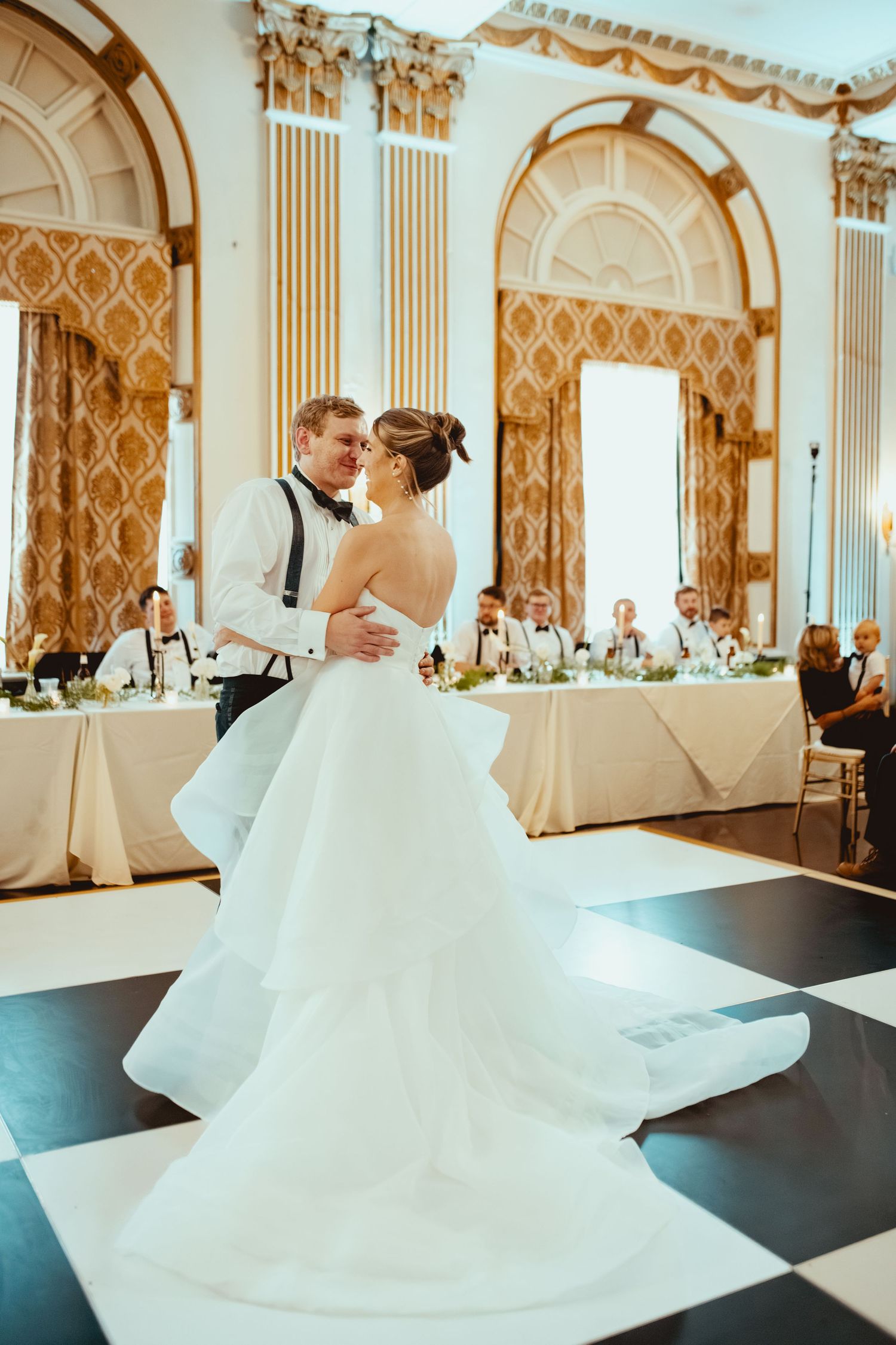 Newlyweds share their first dance in an elegant ballroom with white columns and golden decor.