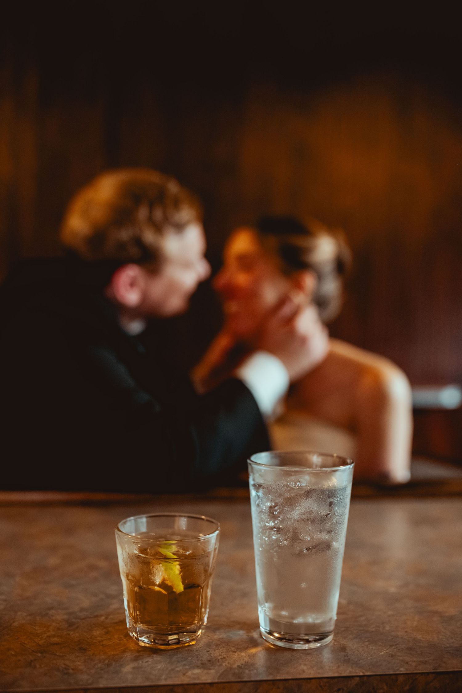 Three glasses on a rustic wooden bar counter showing a whiskey cocktail, water, and ice in dim lighting.
