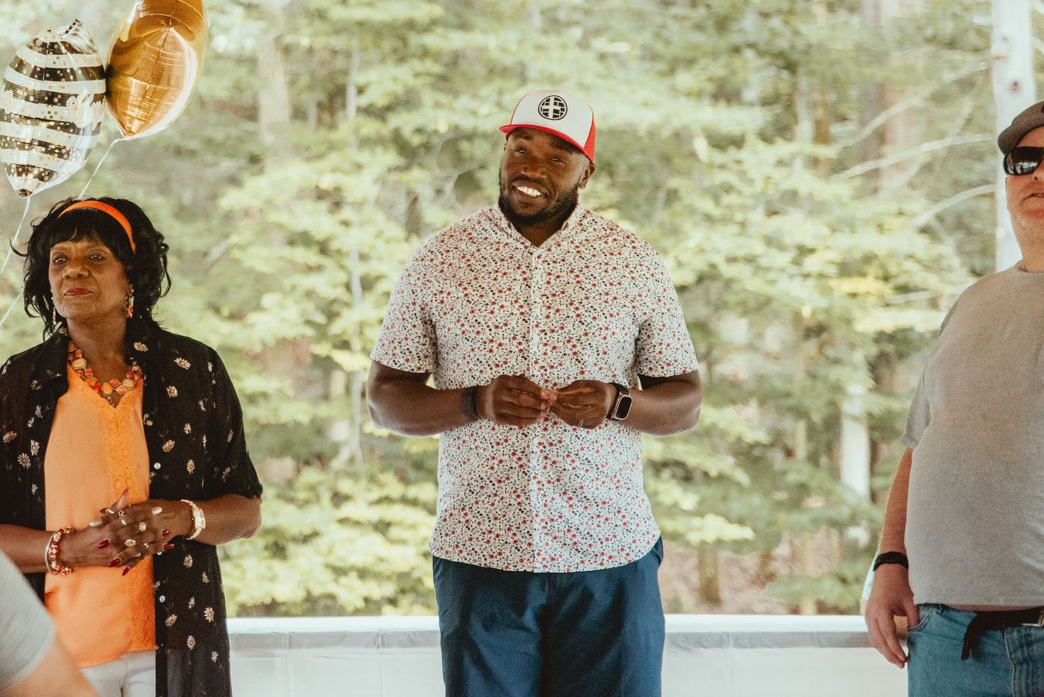 A group of people gather outdoors in casual attire for a social event in a wooded setting.