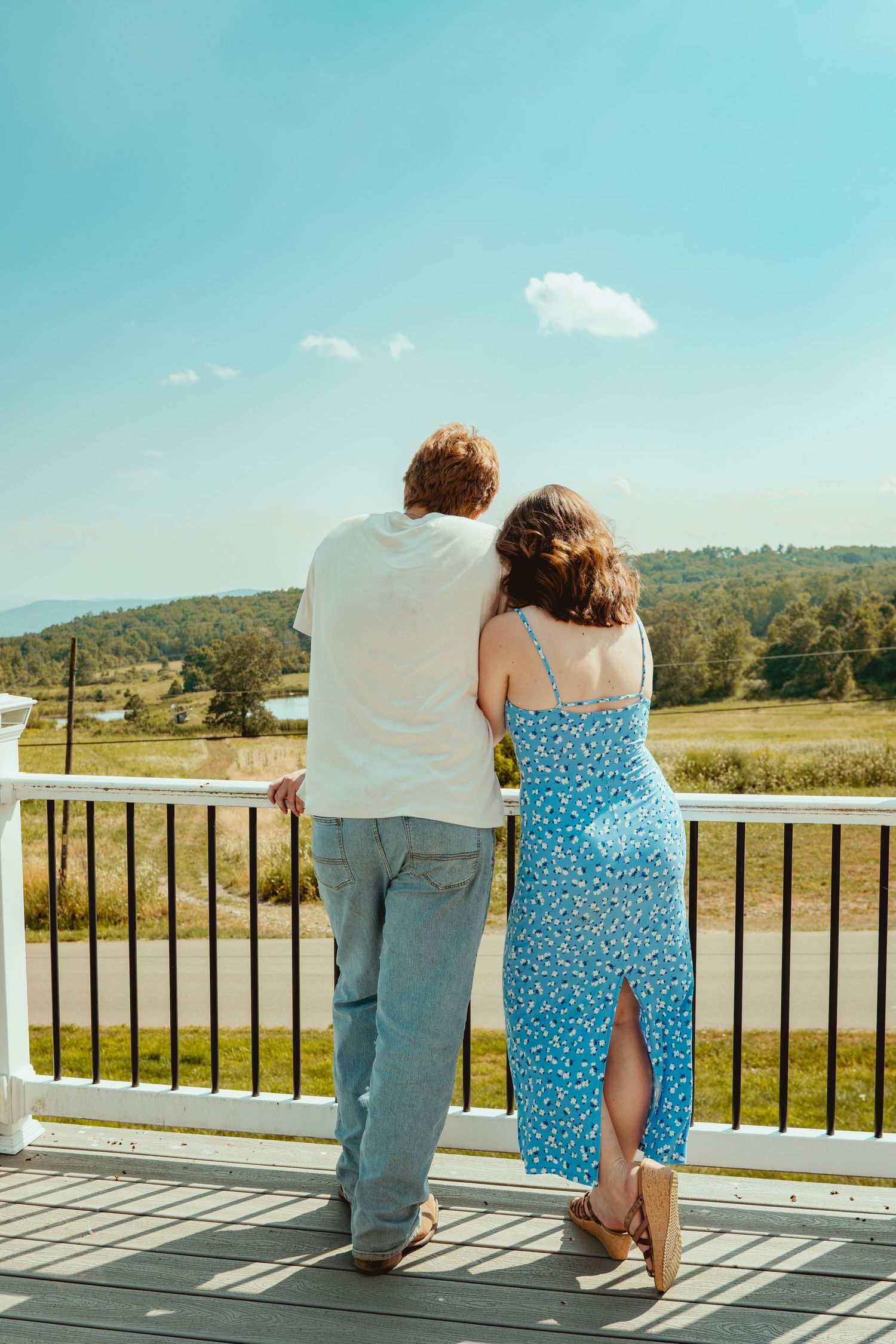 A couple stands together on a sunny deck overlooking scenic countryside views with mountains in the distance.