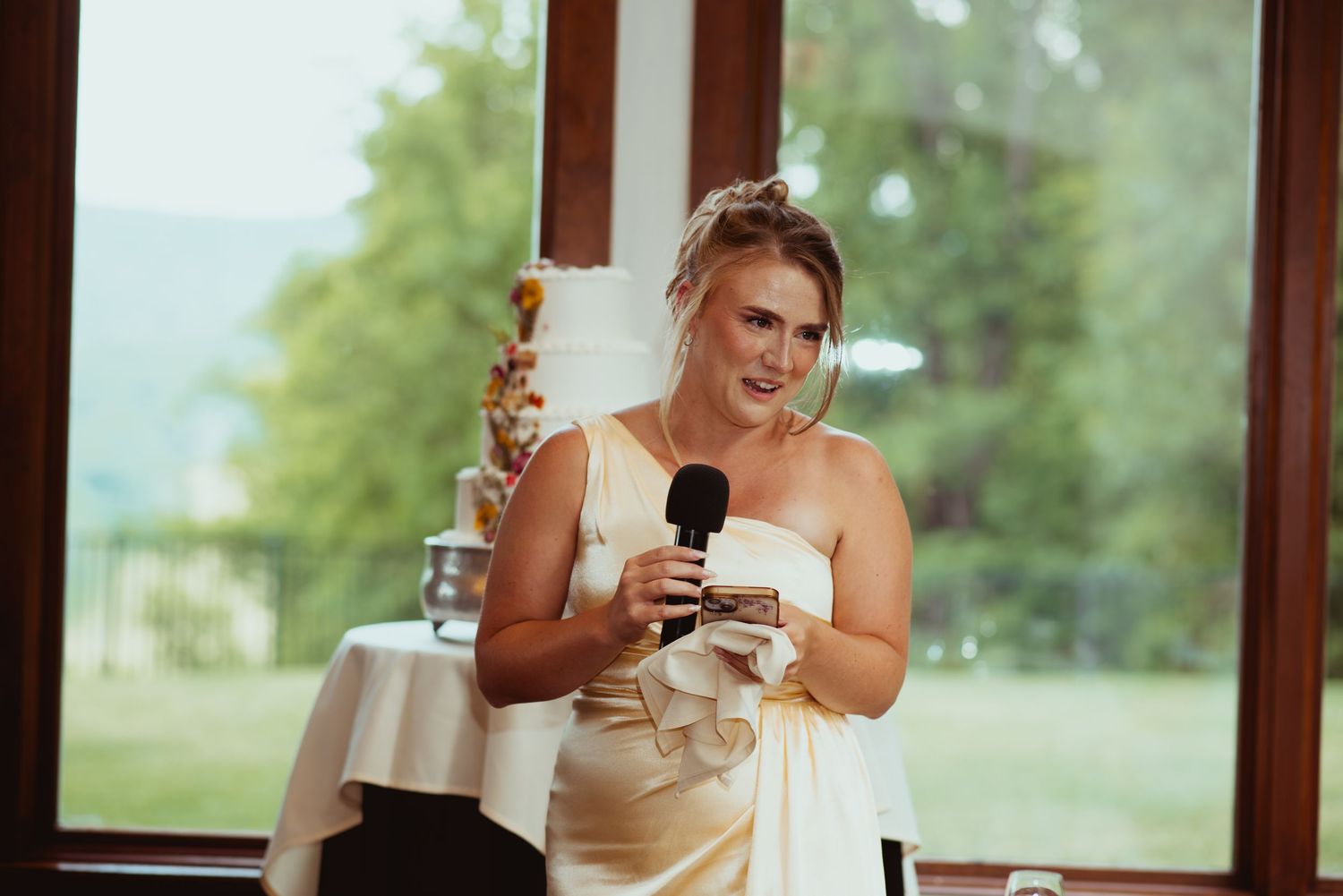 Bridesmaid in ivory one-shoulder gown delivers heartfelt speech with microphone at wedding reception.