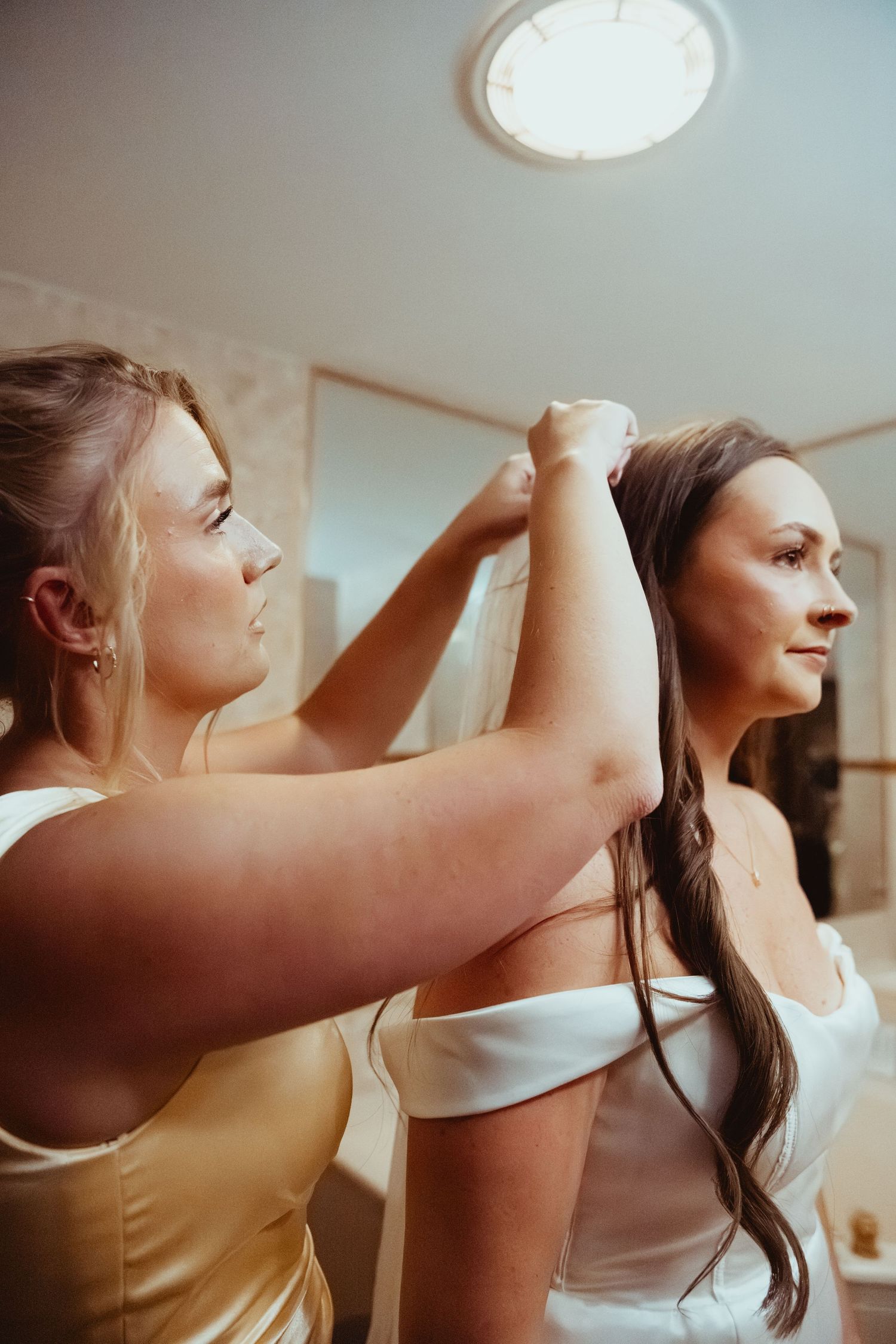 Bridesmaid helping bride put on veil while getting ready in a bright bathroom before the wedding ceremony.