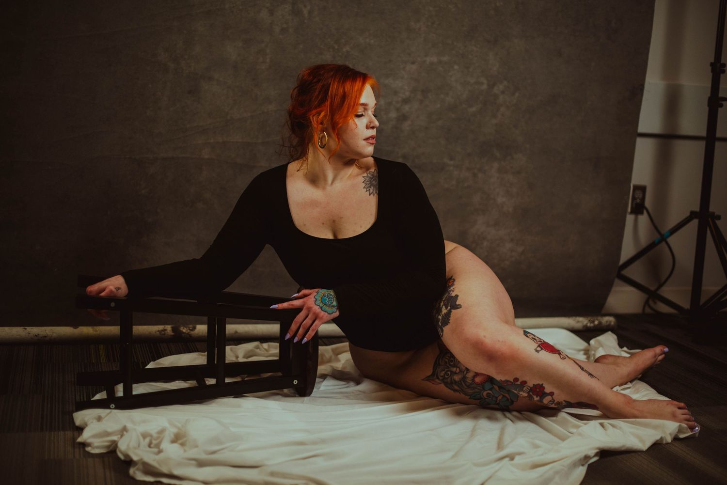 A curvy redhead woman with tattoos poses in a black bodysuit on a white sheet in a moody studio setting.