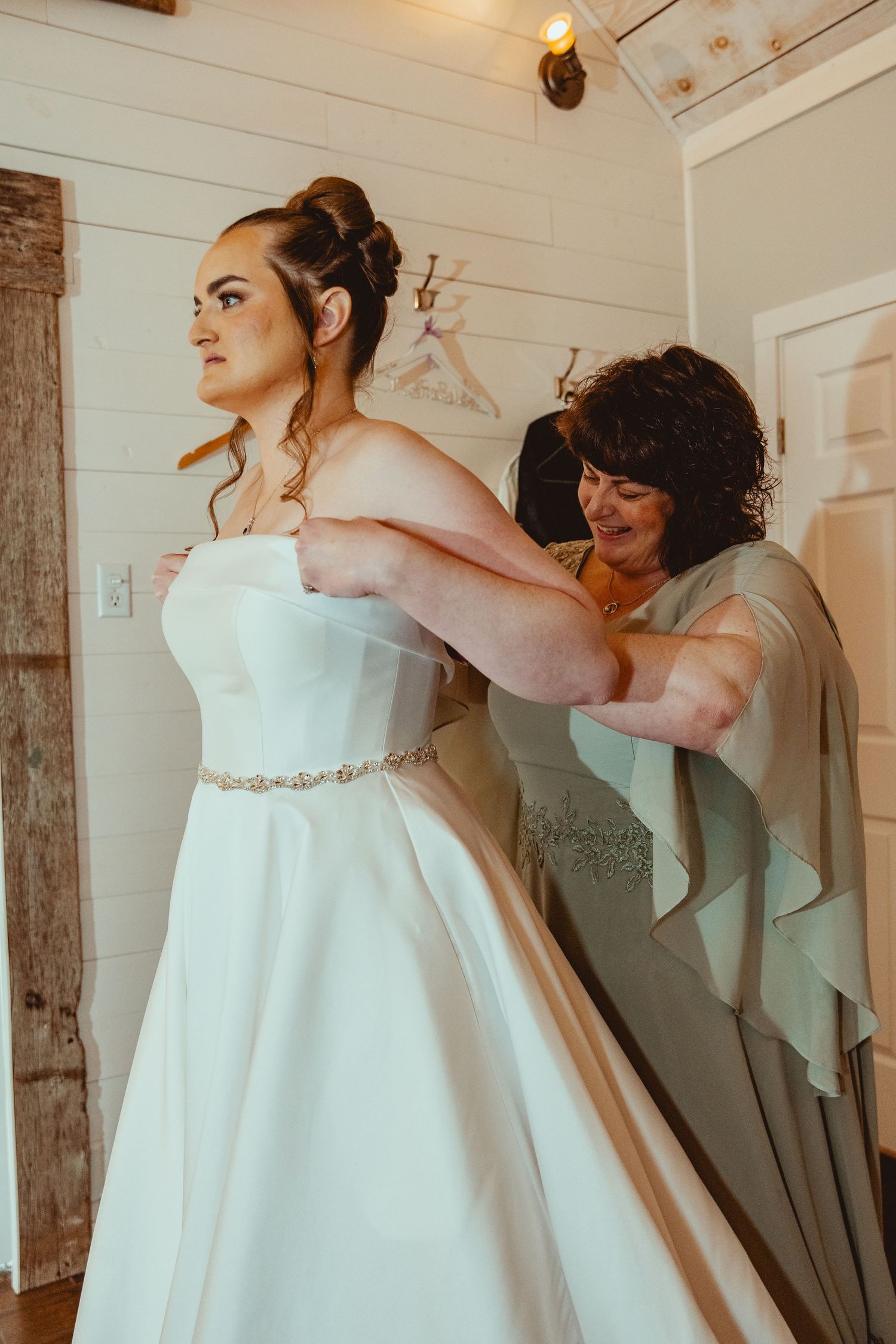 A bride in a white strapless gown gets her dress fastened by a woman in sage green at a rustic wedding venue.