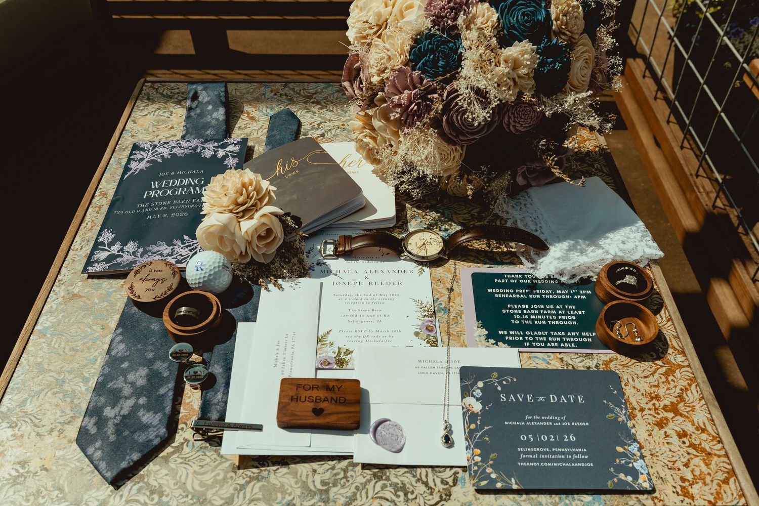 Elegant wedding flat lay featuring navy save-the-date cards, floral invitations, a silk tie, rings, and a lush bouquet on a gold table.