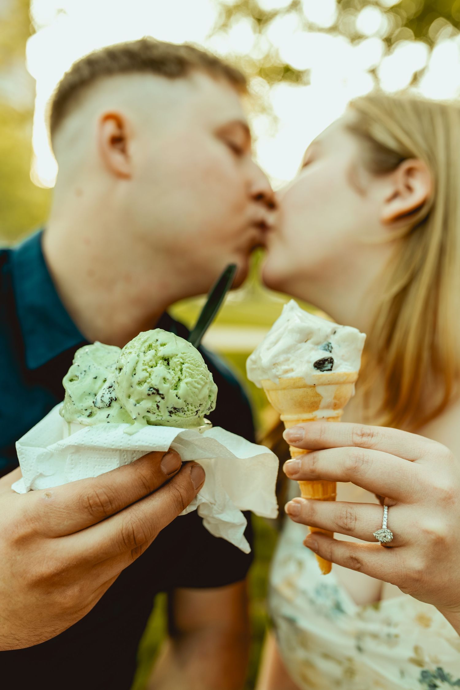 Engaged couple kissing while holding ice cream cones outdoors, woman wearing a diamond engagement ring.