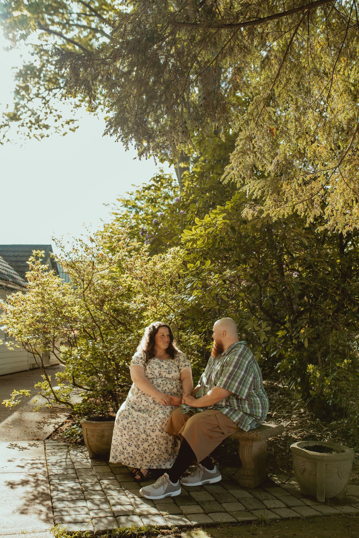 Couple sitting together on a garden bench surrounded by lush green trees in warm golden sunlight.