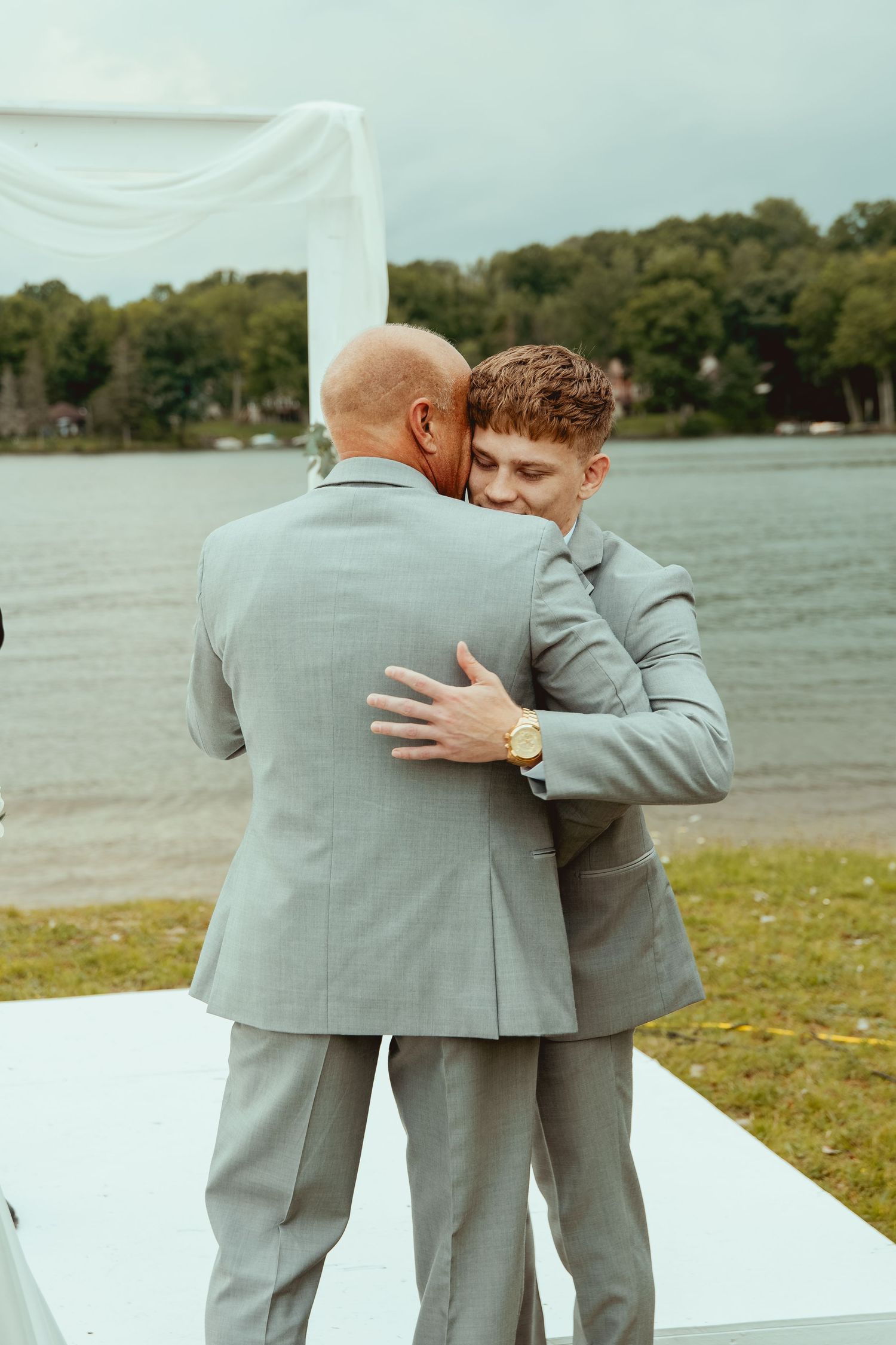 Two groomsmen in gray suits sharing an emotional hug at outdoor lakeside wedding ceremony.
