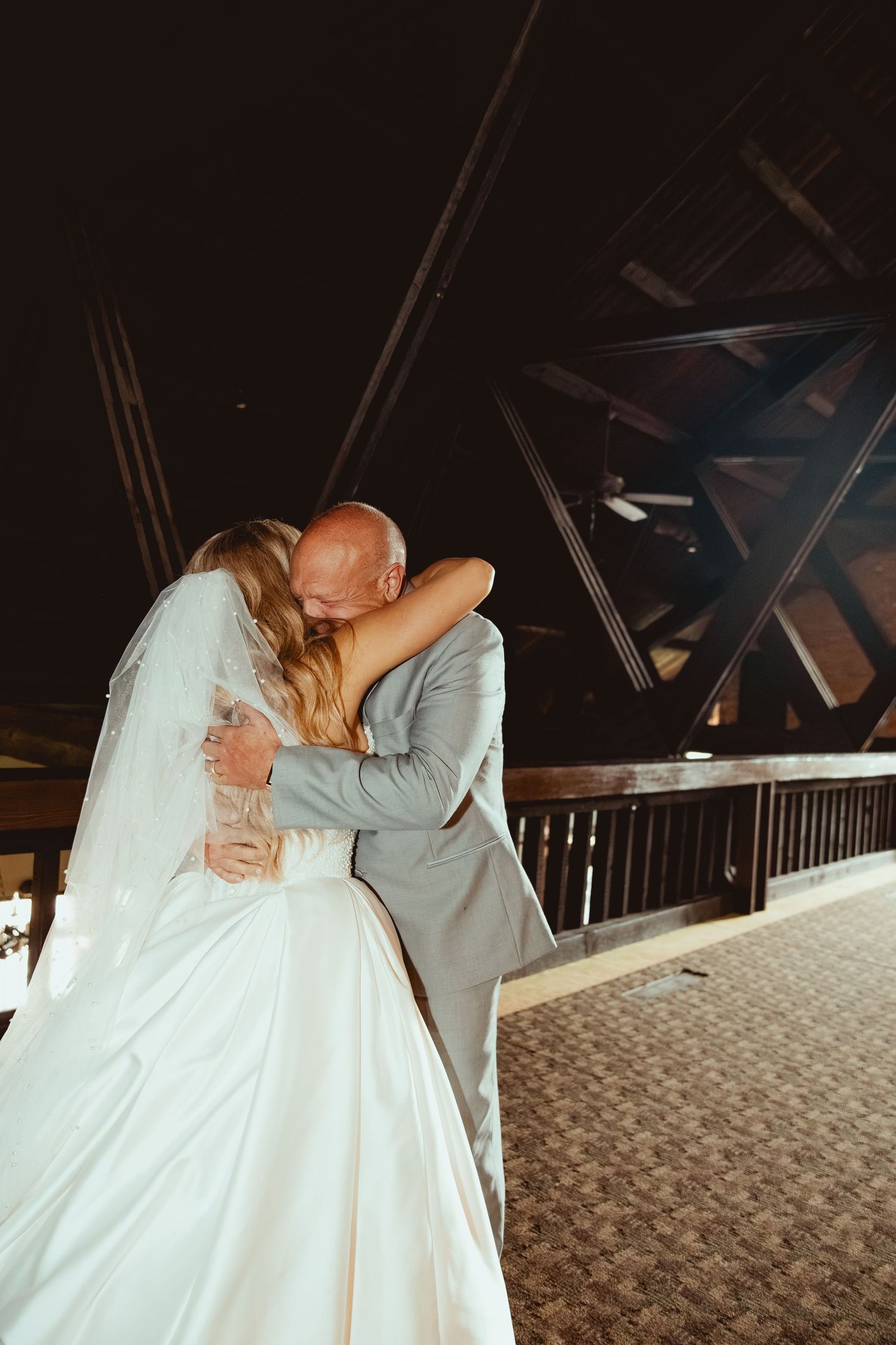 Bride and father share emotional hug during wedding dance under dramatic dark ceiling with steel beams.