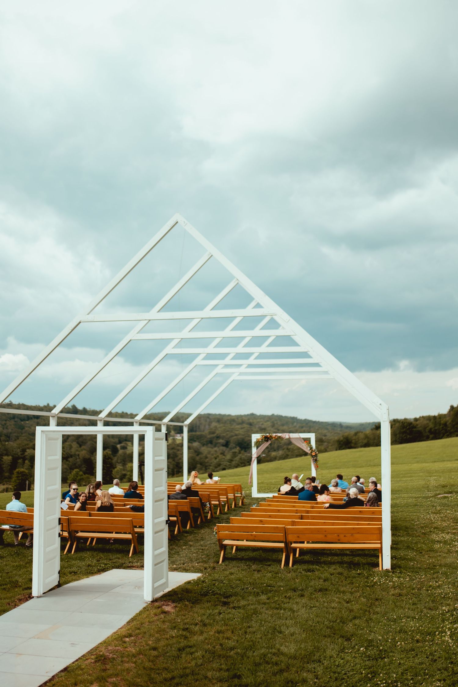 Outdoor wedding ceremony setup with white geometric arch, wooden pews, and rolling green hills under cloudy skies.